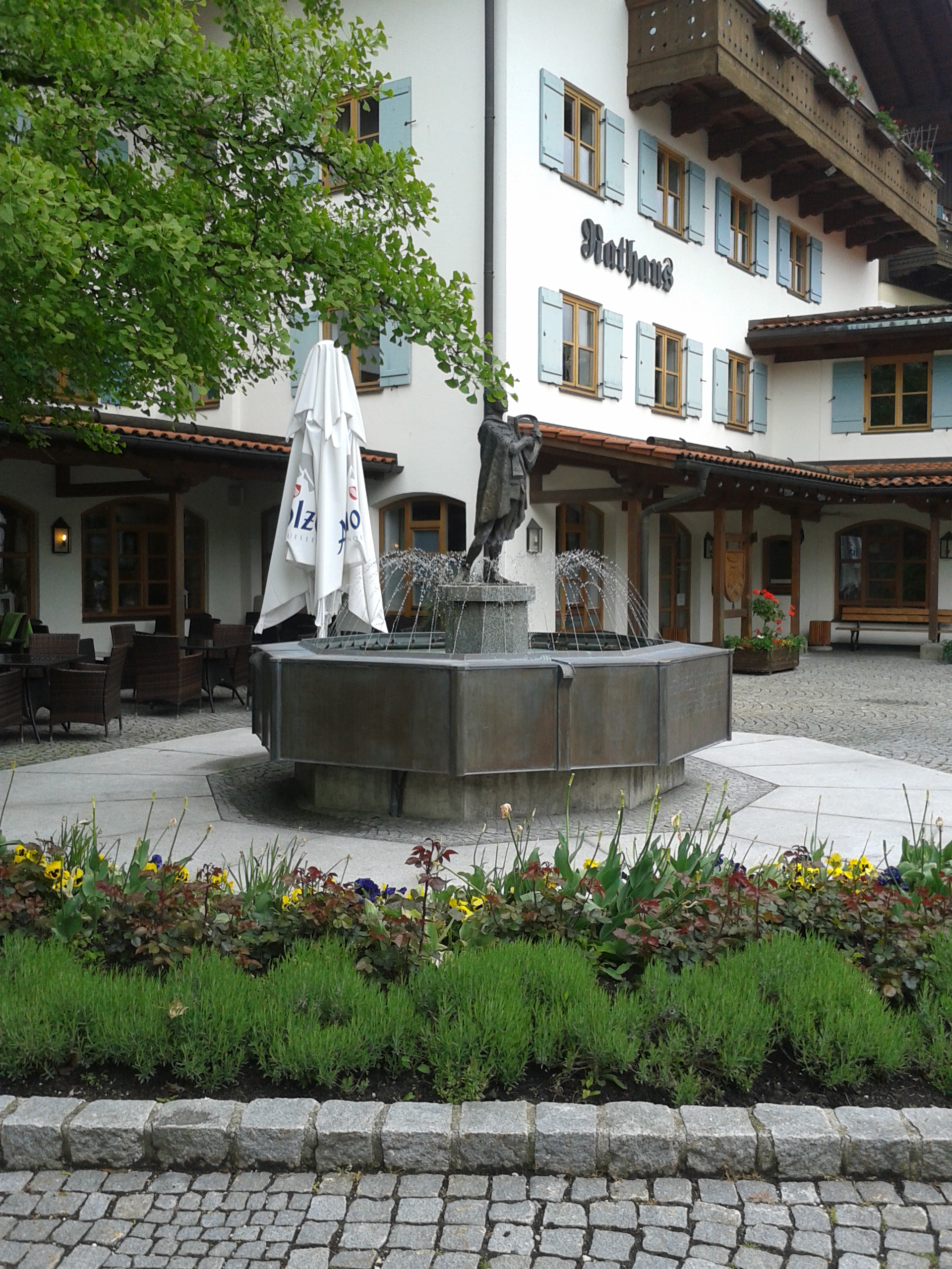 Man sieht einen Weg, daran anschließend ein Blumenbeet. Hinter dem Beet erstreckt sich ein sechseckiger Brunnen, aus dem eine männliche Steinfigur herausragt. Sowohl auf dem gepflasterten Weg, als auch auf dem Platz um den Brunnen herum, werden an Markttagen Stände aufgebaut. Im Hintergrund zeigt sich das Rathaus.